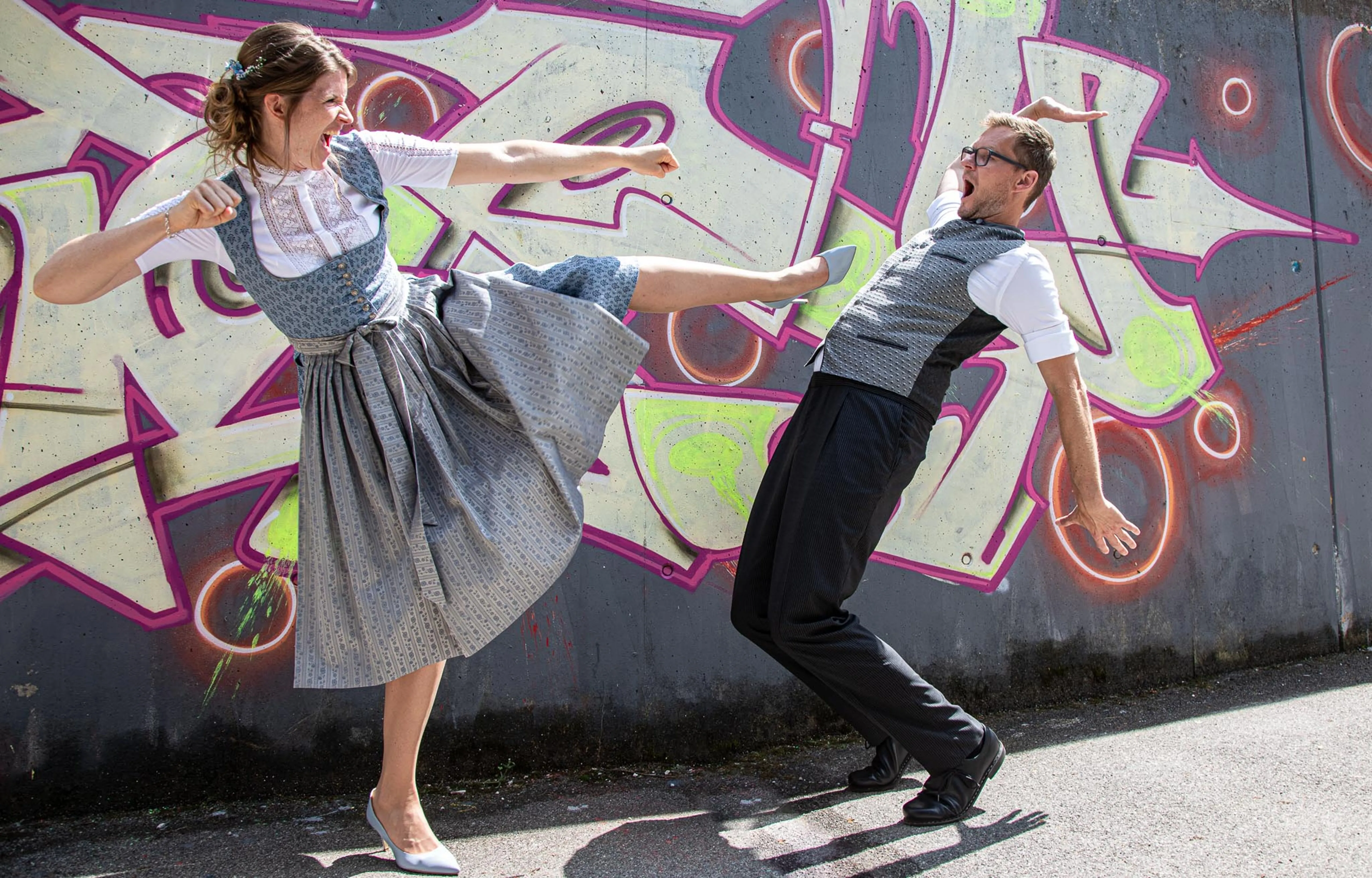 Paar in bayrischer Tracht in verspielter Pose vor bunter Graffitiwand